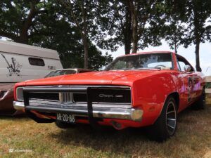 Red vintage Dodge Charger on a grassy lot, front grille and bumper visible, license plate AR-20-88 with a white trailer and trees in the background.