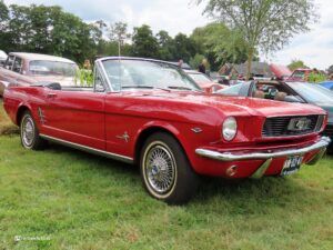Red Ford Mustang convertible on a grassy field at a vintage car show, with other classic cars and onlookers in the background.