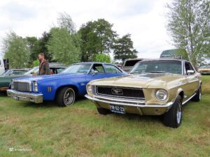 Blue classic car with a man standing beside it at a grassy car show; other vintage cars nearby.