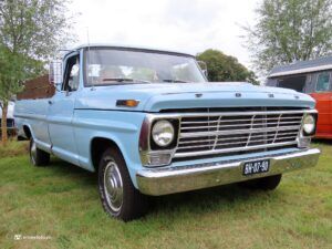 Front-left view of a light blue vintage Ford pickup with a chrome grille parked on grass, with a gray and orange camper van in the background on the right.