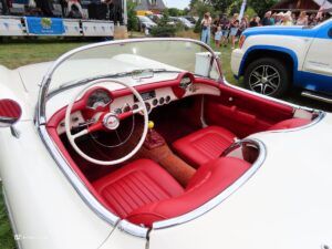Interior of a white vintage convertible with red leather seats and white steering wheel, chrome trim, and classic dashboard details visible, at an outdoor event.