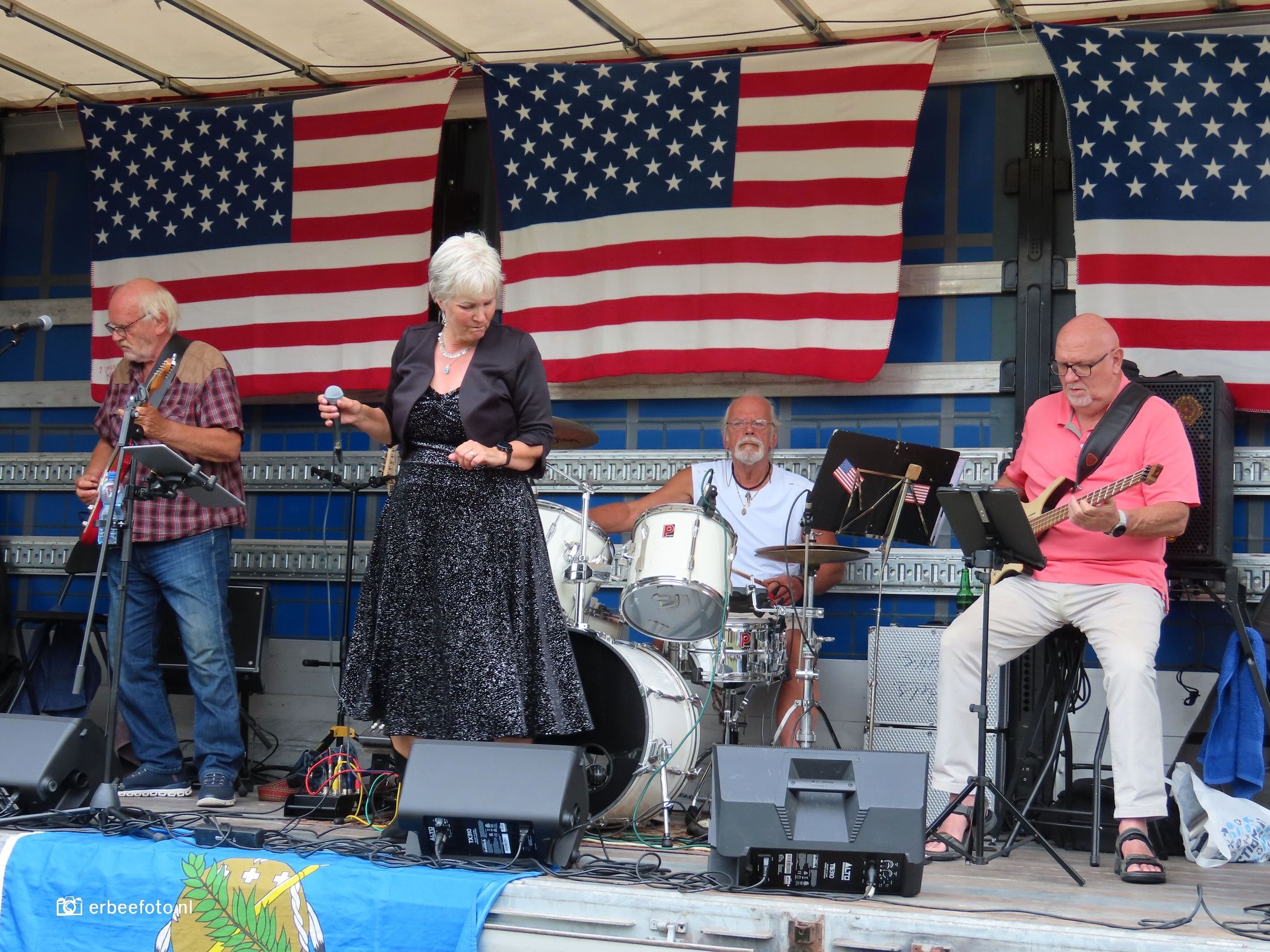 American Car Event - De Wilp 23 – Westerkwartier|NU Live band performing on stage with three musicians and large American flag banners as backdrop