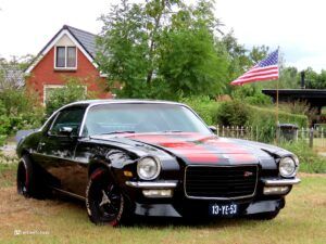 Front view of a black vintage Chevrolet Camaro Z28 parked on a grassy lawn in front of a red-brick house, with a large tree and an American flag in the background.