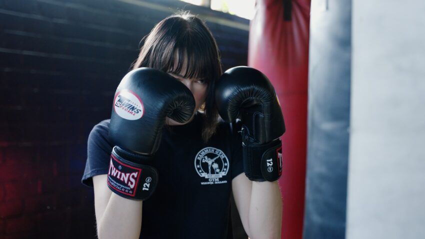 Person wearing black boxing gloves in a gym, hands up in a guarding stance.